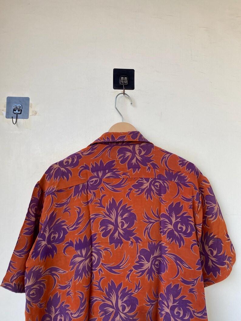 May include: Orange short-sleeved shirt with a purple floral pattern. The shirt is hanging on a wooden hanger. The floral design is a repeating pattern of large, stylized flowers.