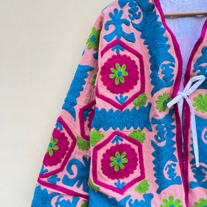 Puede incluir: Una chaqueta rosa con patrones florales bordados en azul, verde y rosa. La chaqueta tiene un cierre de corbata en la parte delantera.