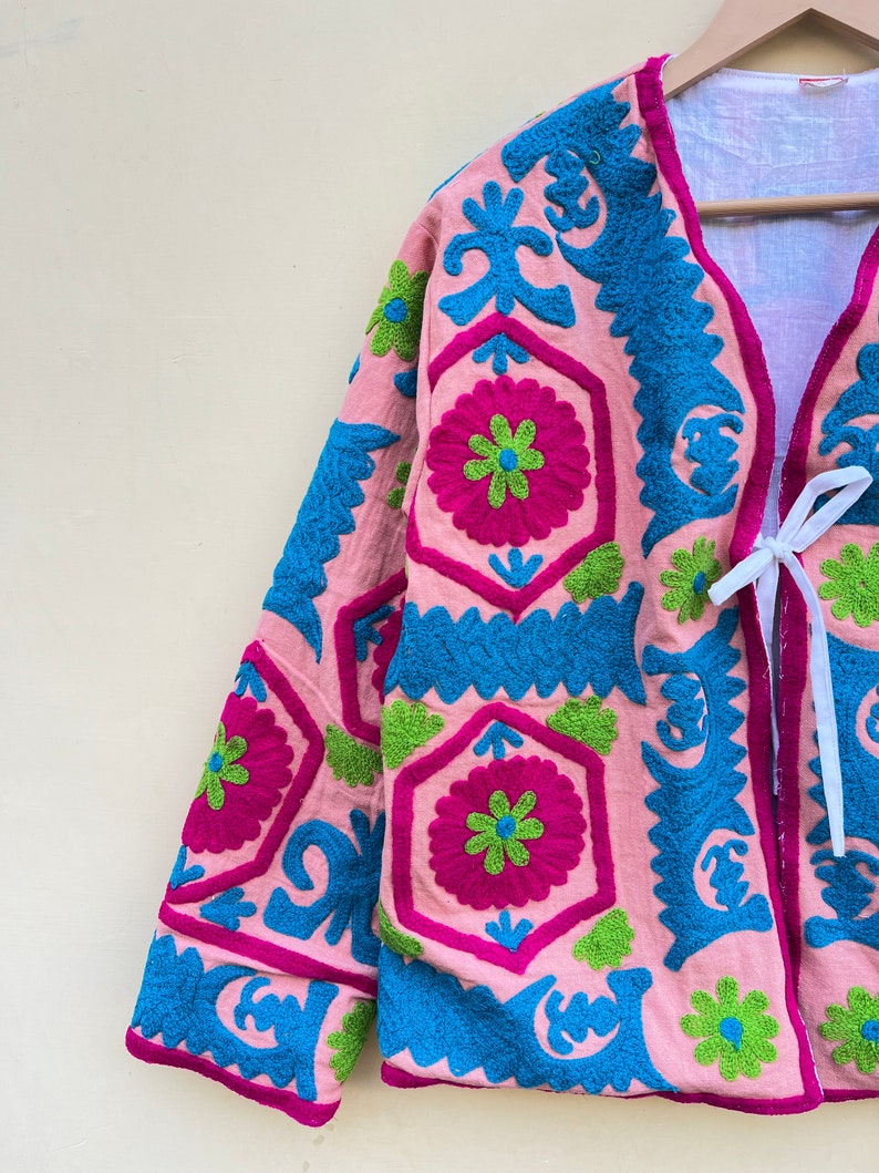 Puede incluir: Una chaqueta rosa con patrones florales bordados en azul, verde y rosa. La chaqueta tiene un cierre de corbata en la parte delantera.