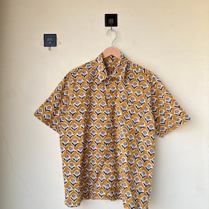 May include: Short-sleeved button-down shirt with a repeating floral pattern in mustard yellow, navy blue, and white. The shirt is hanging on a wooden hanger against a light yellow wall. The shirt's collar is visible.