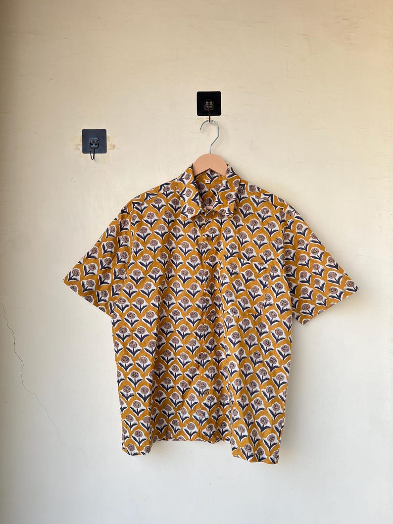 May include: Short-sleeved button-down shirt with a repeating floral pattern in mustard yellow, navy blue, and white. The shirt is hanging on a wooden hanger against a light yellow wall. The shirt's collar is visible.