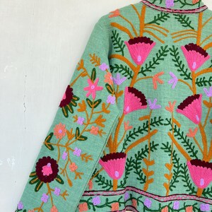 May include: A mint green jacket with embroidered floral details in shades of pink, orange, and purple. The jacket has a stand-up collar and long sleeves.