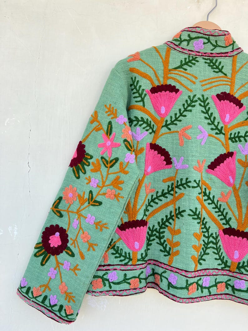 May include: A mint green jacket with embroidered floral details in shades of pink, orange, and purple. The jacket has a stand-up collar and long sleeves.