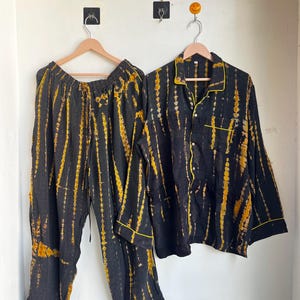 Może przedstawiać: Czarny zestaw piżamy z ż&oacute;łtym wzorem tie-dye. Zestaw zawiera koszulę zapinaną na guziki z ż&oacute;łtymi wykończeniami i pasujące spodnie. Piżamy wiszą na drewnianych wieszakach na białej ścianie.