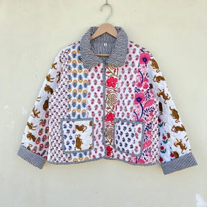 Può includere: Una giacca trapuntata bianca con un design a patchwork che presenta vari motivi floreali e animali. La giacca ha un colletto, maniche lunghe e due tasche a toppa.