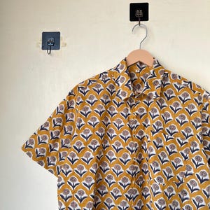May include: Mustard yellow short-sleeved shirt with a repeating floral pattern in white, navy blue, and brown. The shirt is hanging on a wooden hanger against a neutral wall. The floral design is evenly spaced across the fabric.