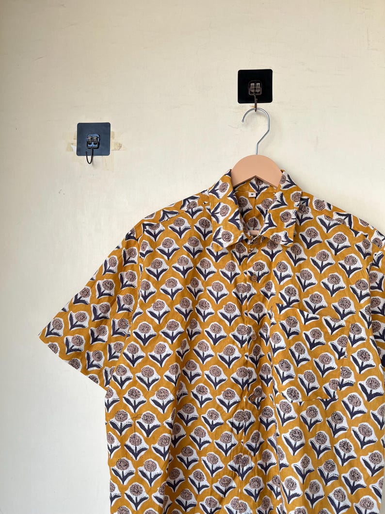 May include: Mustard yellow short-sleeved shirt with a repeating floral pattern in white, navy blue, and brown. The shirt is hanging on a wooden hanger against a neutral wall. The floral design is evenly spaced across the fabric.