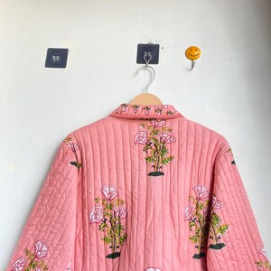 Pu&ograve; includere: Giacca trapuntata rosa con motivo floreale. La giacca presenta un motivo ripetuto di fiori rosa e bianchi con foglie verdi e steli neri. La giacca &egrave; appesa a un appendiabiti in legno.