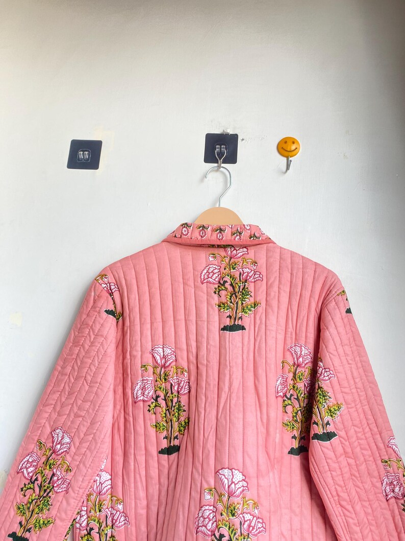 Pu&ograve; includere: Giacca trapuntata rosa con motivo floreale. La giacca presenta un motivo ripetuto di fiori rosa e bianchi con foglie verdi e steli neri. La giacca &egrave; appesa a un appendiabiti in legno.