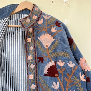 May include: A blue velvet jacket with intricate floral embroidery in shades of pink, brown, and red. The jacket has a striped lining and a mandarin collar.