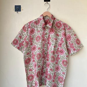 May include: A short-sleeved button-down shirt with a floral pattern. The shirt features a white background with a repeating pattern of pink flowers and green leaves. The shirt is hanging on a wooden hanger.