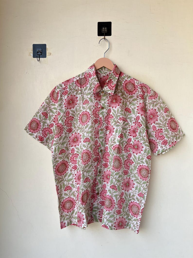 May include: A short-sleeved button-down shirt with a floral pattern. The shirt features a white background with a repeating pattern of pink flowers and green leaves. The shirt is hanging on a wooden hanger.
