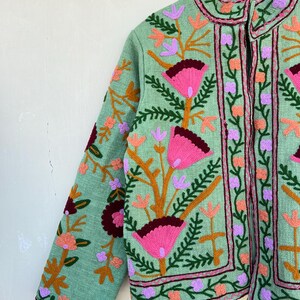May include: A green embroidered jacket with a floral pattern in shades of pink, purple, orange, and brown. The jacket has a mandarin collar and a front zipper closure.