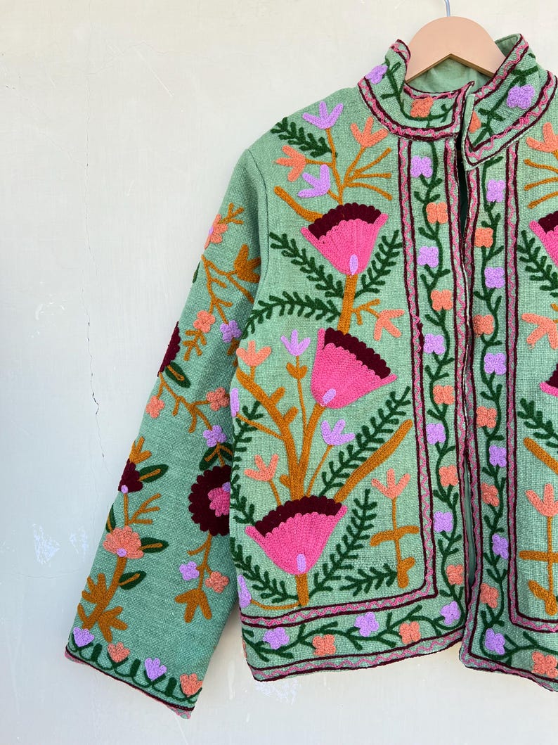 May include: A green embroidered jacket with a floral pattern in shades of pink, purple, orange, and brown. The jacket has a mandarin collar and a front zipper closure.