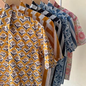 May include: A row of short-sleeved shirts in various patterns and colors hang on a white rack. The shirts feature floral, striped, and geometric designs in shades of yellow, blue, black, and pink. The shirts are made of a lightweight fabric.