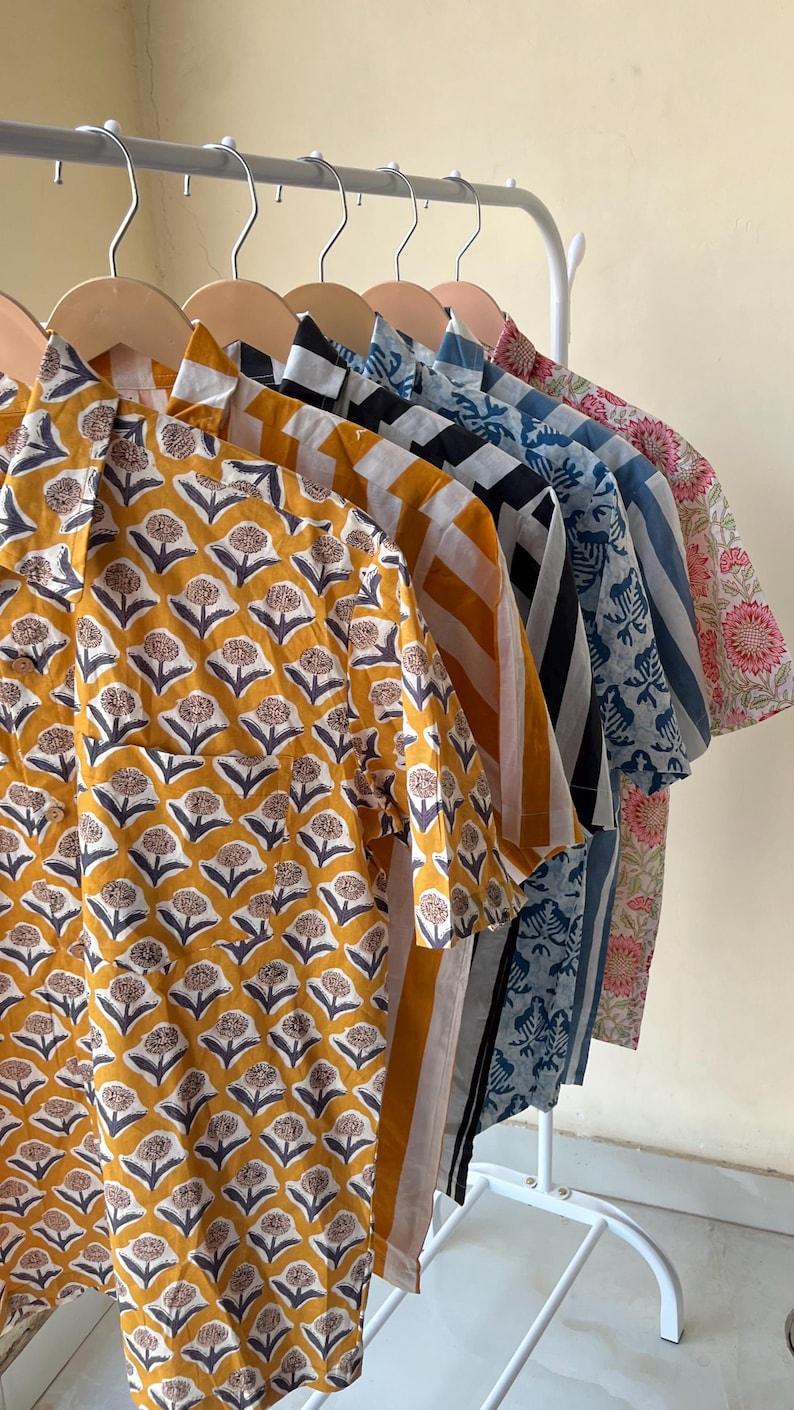 May include: A row of short-sleeved shirts in various patterns and colors hang on a white rack. The shirts feature floral, striped, and geometric designs in shades of yellow, blue, black, and pink. The shirts are made of a lightweight fabric.