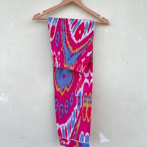 May include: Pink, blue, and gold patterned pants hanging on a wooden hanger. The pants have a white trim around the bottom.