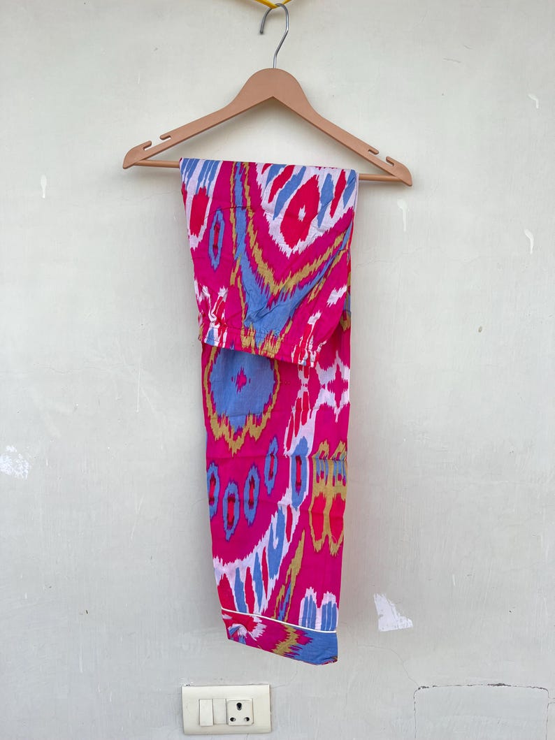 May include: Pink, blue, and gold patterned pants hanging on a wooden hanger. The pants have a white trim around the bottom.