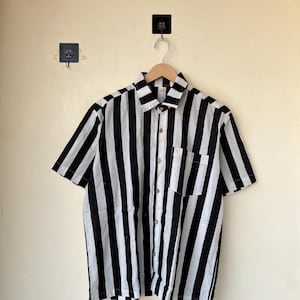 May include: A short-sleeved shirt with bold black and white vertical stripes. The shirt has a collared neckline, a single chest pocket, and wooden buttons. It is hanging on a wooden hanger against a light-colored wall.