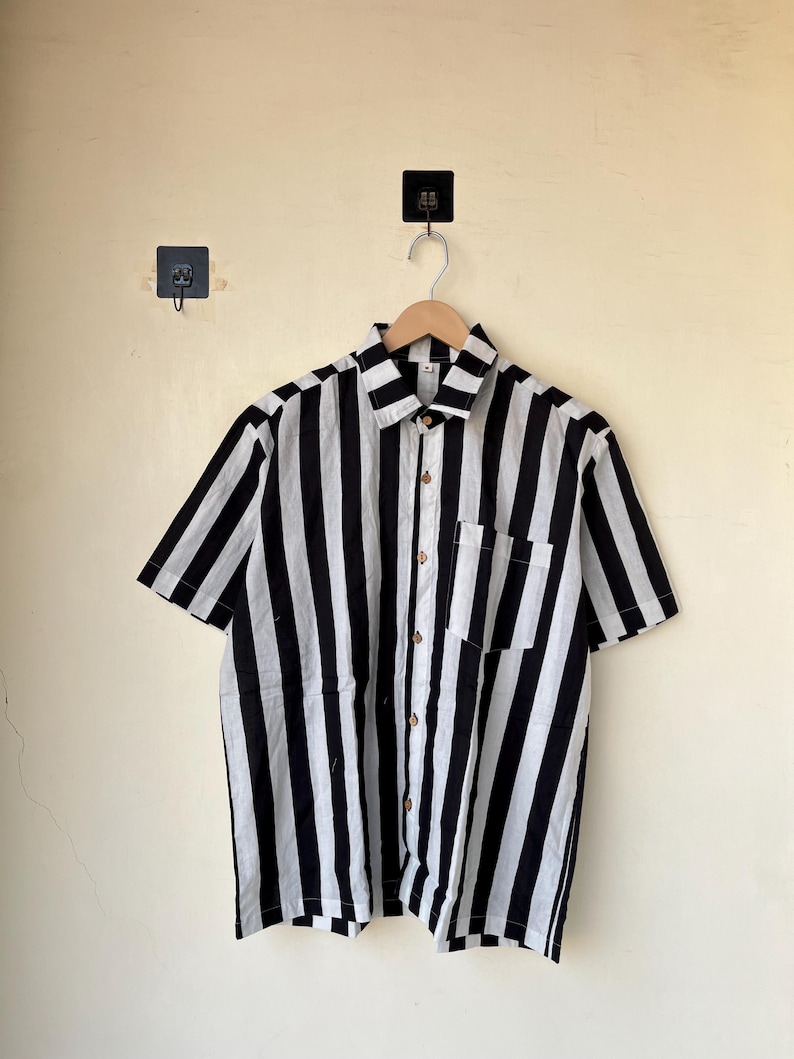 May include: A short-sleeved shirt with bold black and white vertical stripes. The shirt has a collared neckline, a single chest pocket, and wooden buttons. It is hanging on a wooden hanger against a light-colored wall.