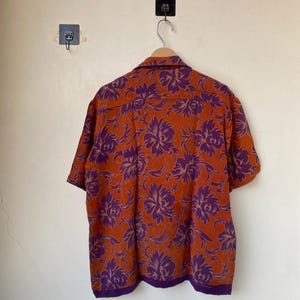 May include: Short-sleeved button-down shirt with a vibrant floral pattern. The shirt features a burnt orange background with large purple floral designs. The collar and hem are trimmed with a darker purple fabric. The shirt is hanging on a wooden hanger.