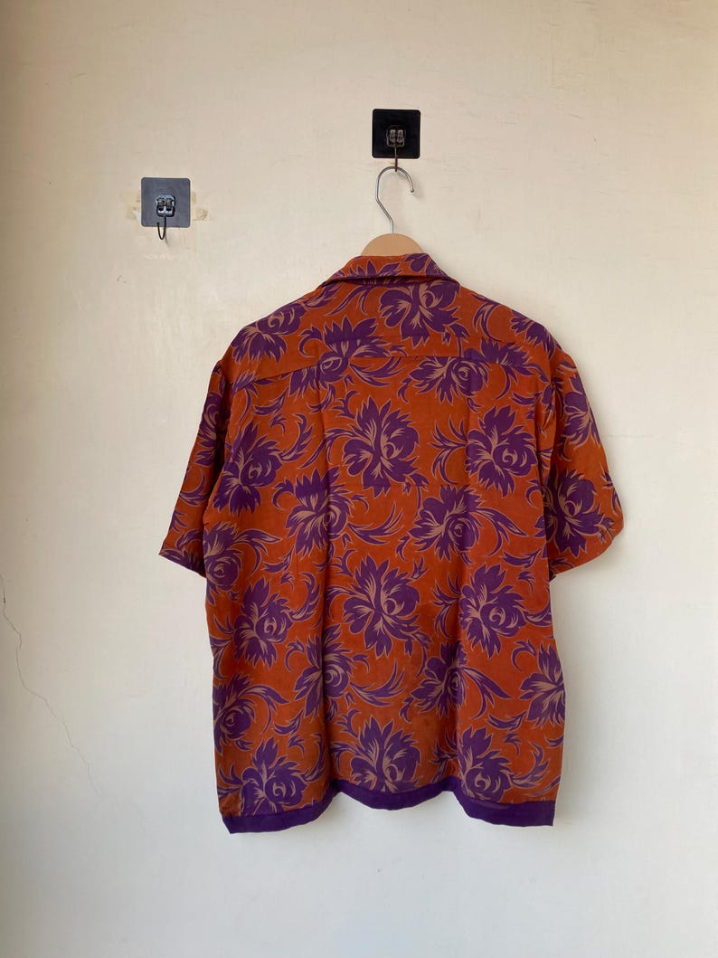 May include: Short-sleeved button-down shirt with a vibrant floral pattern. The shirt features a burnt orange background with large purple floral designs. The collar and hem are trimmed with a darker purple fabric. The shirt is hanging on a wooden hanger.