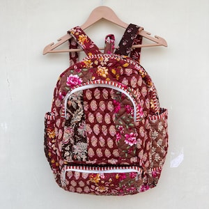 Op de afbeelding: Een gewatteerde rugzak met een bloemenpatroon in tinten rood, bruin en geel. De rugzak heeft een ritsvak aan de voorkant en een witte rand rond de randen.