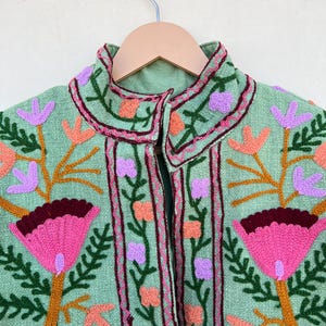 May include: A green embroidered jacket with pink, orange, and purple floral designs. The jacket has a high collar and a button closure.