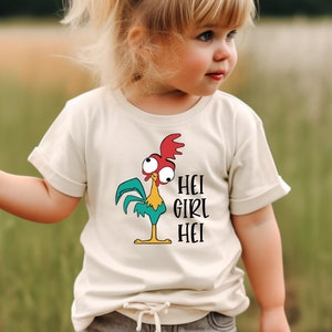 Pu&ograve; includere: Una maglietta beige chiaro per bambini con un gallo dei cartoni animati con accenti rossi, blu e gialli. Il testo "HEI GIRL HEI" &egrave; stampato in nero. Il bambino indossa pantaloncini grigi e indica verso sinistra.