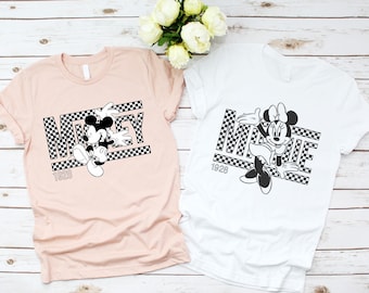 Può includere: Due t-shirt con un designo a scacchi bianco e nero con Mickey Mouse e Minnie Mouse. La maglietta di Mickey Mouse è rosa e quella di Minnie Mouse è bianca. Entrambe le magliette hanno il testo "1928" e il nome del personaggio in una scatola a scacchi bianco e nero.