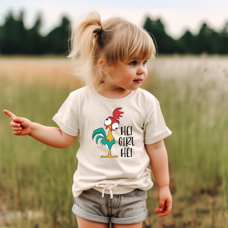 Pu&ograve; includere: Una maglietta beige chiaro per bambini con un gallo dei cartoni animati con accenti rossi, blu e gialli. Il testo "HEI GIRL HEI" &egrave; stampato in nero. Il bambino indossa pantaloncini grigi e indica verso sinistra.