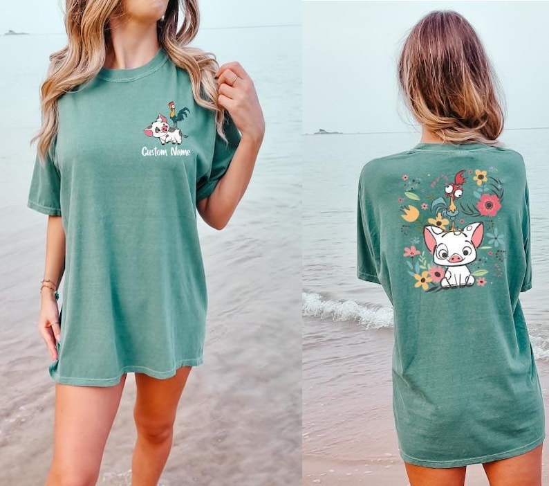 May include: A sage green short-sleeve t-shirt featuring a small graphic on the front chest of a pig and a rooster with the text Custom Name. The back of the shirt displays a larger, colorful illustration of the same pig and rooster among flowers.