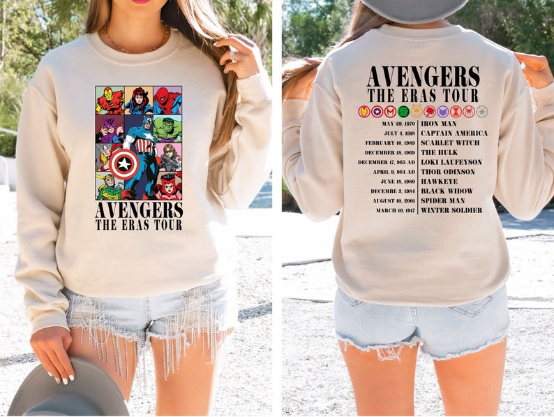 May include: A beige sweatshirt with a graphic print of the Avengers characters and the text "Avengers The Eras Tour". The back of the sweatshirt has a list of the Avengers characters and their first appearances.