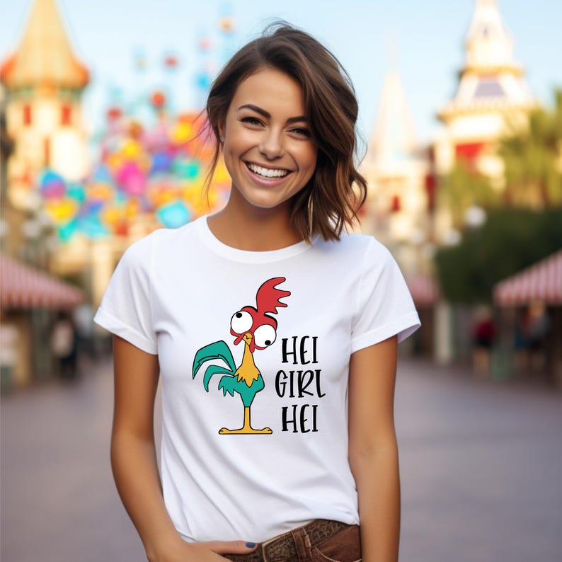 Pu&ograve; includere: T-shirt bianca con un disegno di gallo dei cartoni animati con accenti rossi, verdi e gialli. Il testo "HEI GIRL HEI" &egrave; stampato in nero. La maglietta ha un girocollo e maniche corte. Lo sfondo &egrave; sfocato.
