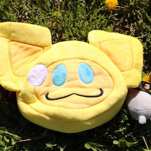 May include: A plush yellow character with large ears, a smiling face with blue eyes, and a small gray plush figure with a patterned collar. The items are set on a bed of green grass with yellow dandelions.