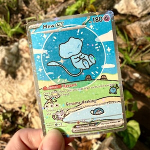 May include: A holographic Mew ex Pok&eacute;mon trading card. The card features a light blue Mew character in a circular design with a starry background. The card has text including "Mew ex", "HP 180", and attack descriptions.