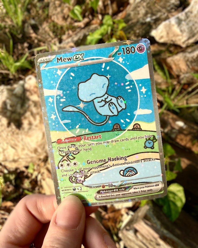 May include: A holographic Mew ex Pok&eacute;mon trading card. The card features a light blue Mew character in a circular design with a starry background. The card has text including "Mew ex", "HP 180", and attack descriptions.