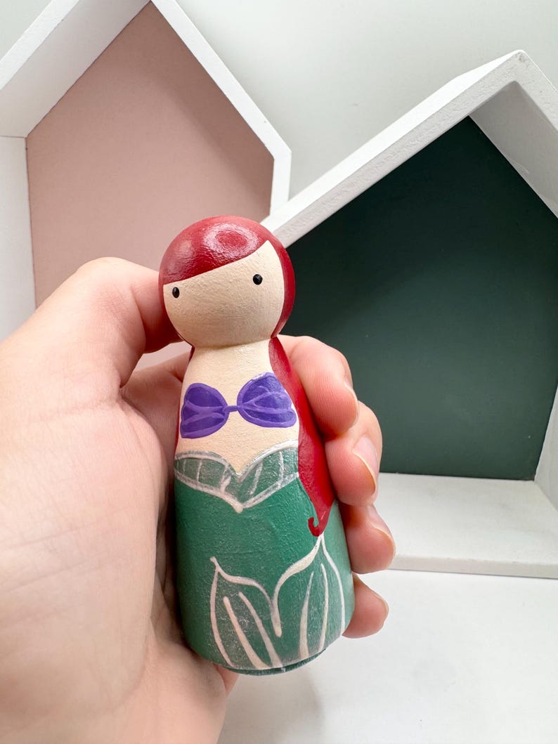 Puede incluir: Mu&ntilde;eca de madera pintada a mano que representa un personaje con cabello rojo, un top morado y una falda verde. La mu&ntilde;eca se sostiene contra un fondo de formas geom&eacute;tricas blancas y verdes, lo que sugiere un art&iacute;culo decorativo o de colecci&oacute;n.