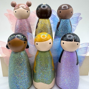 Puede incluir: Un conjunto de seis muñecos de madera con caras pintadas y vestidos brillantes en varios colores, incluyendo rosa, amarillo, azul, verde y morado. Cada muñeco tiene alas de tela iridiscente transparente en la espalda y peinados distintos.