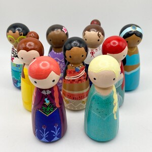 Puede incluir: Una colecci&oacute;n de mu&ntilde;ecas de madera pintadas a mano, cada una representando un personaje diferente con rasgos faciales, peinados y atuendos coloridos &uacute;nicos. Las mu&ntilde;ecas est&aacute;n dispuestas en grupo, mostrando una variedad de dise&ntilde;os y temas.