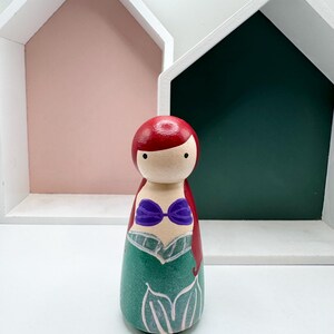 Puede incluir: Mu&ntilde;eca de madera pintada a mano que representa a una sirena. La mu&ntilde;eca tiene pelo rojo, un top de concha p&uacute;rpura y una cola verde. Se encuentra frente a estantes blancos con forma de casa con fondos rosas y verdes.