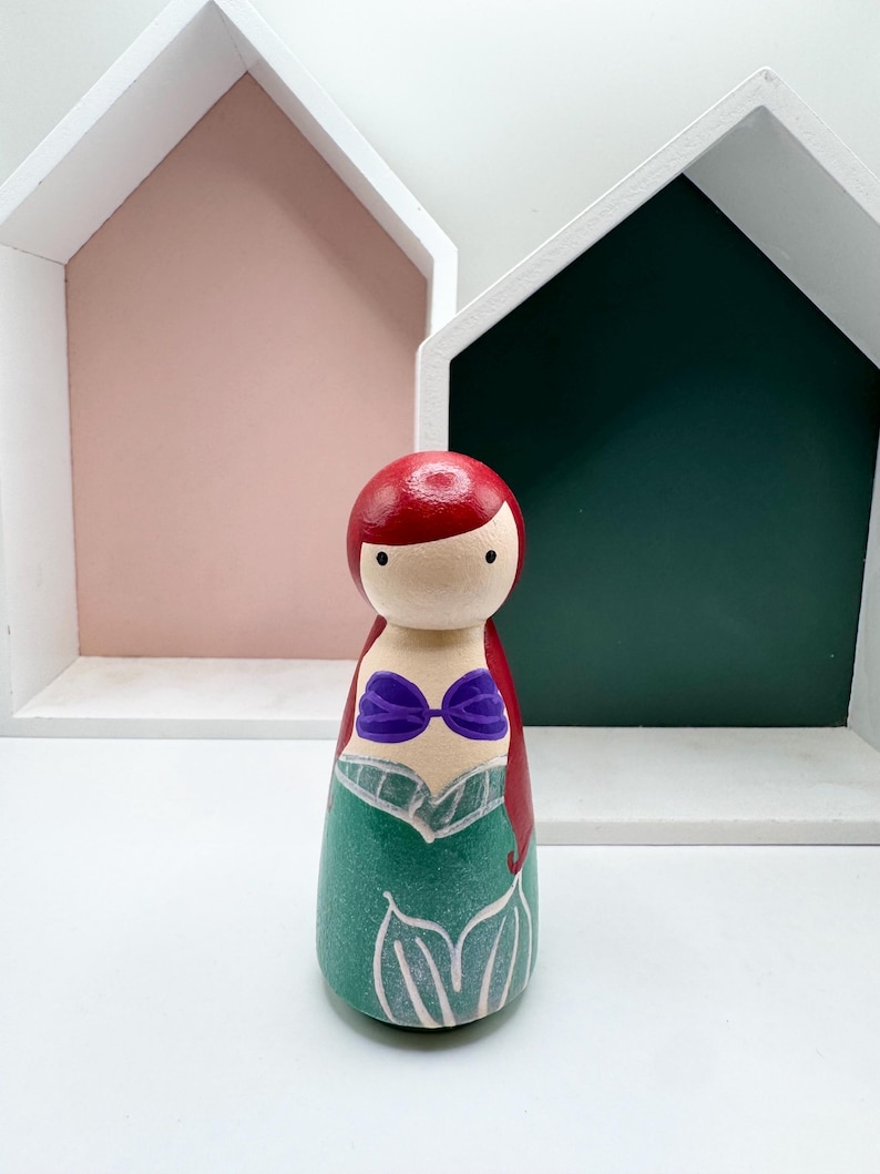 Puede incluir: Mu&ntilde;eca de madera pintada a mano que representa a una sirena. La mu&ntilde;eca tiene pelo rojo, un top de concha p&uacute;rpura y una cola verde. Se encuentra frente a estantes blancos con forma de casa con fondos rosas y verdes.