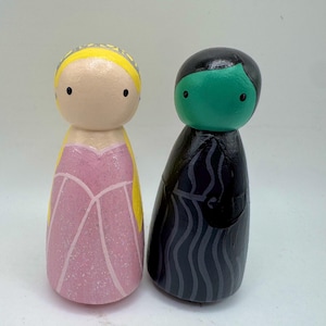 May include: A pair of wooden peg dolls, one dressed in a pink gown with a yellow crown and the other in a black and grey robe with a green face.