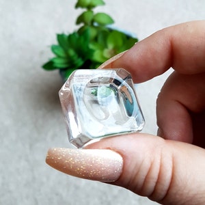 Puede incluir: Un joyero de anillo de vidrio transparente con forma cuadrada. El joyero est&aacute; siendo sostenido por una mano con esmalte de u&ntilde;as rosa claro.