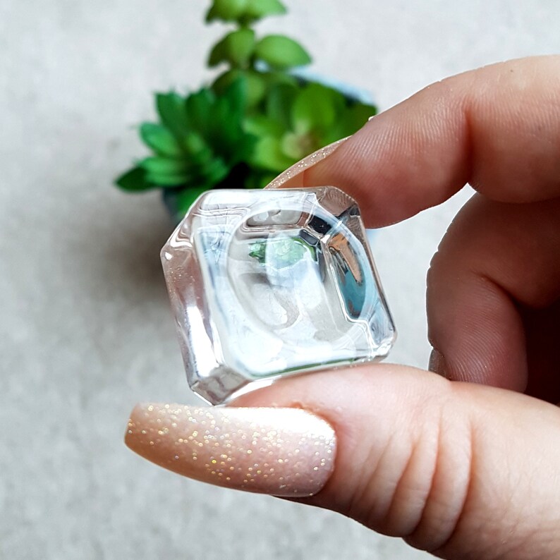 Puede incluir: Un joyero de anillo de vidrio transparente con forma cuadrada. El joyero est&aacute; siendo sostenido por una mano con esmalte de u&ntilde;as rosa claro.