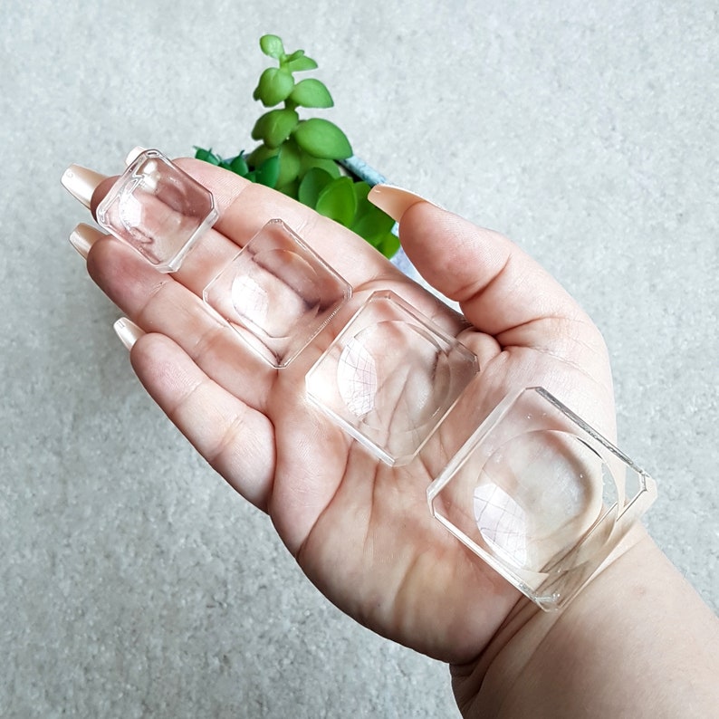 Puede incluir: Cuatro platos cuadrados de pl&aacute;stico transparente para anillos, de diferentes tama&ntilde;os, sostenidos en una mano.