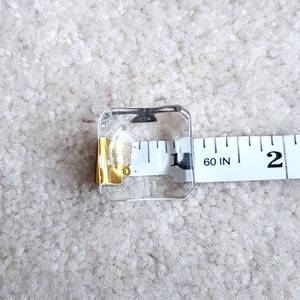 Puede incluir: Un cuadrado de vidrio transparente con una pieza de metal dorado unida a un lado. El cuadrado est&aacute; sobre una cinta m&eacute;trica blanca que dice "60 IN 2".