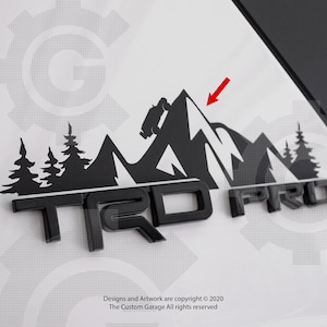 May include: Black and white decal of a mountain range with a silhouette of a truck driving up a mountain. The decal is placed on a black "TRD PRO" emblem.