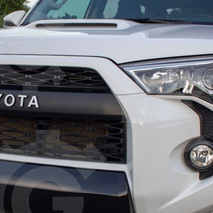 May include: The front end of a white vehicle featuring a black grille with the letters YOTA visible, a hood scoop, clear headlights, and a round fog light. The design includes a black bumper section and a black honeycomb mesh pattern.