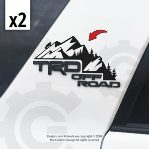 May include: Black TRD Off Road decal featuring a mountain range and trees. The decal includes the text "TRD OFF ROAD" and a small vehicle graphic. The image shows two decals, with a red arrow pointing to one.